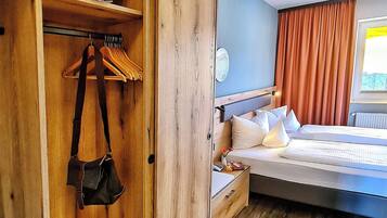 Una habitación de hotel con un armario de madera, una cama con ropa blanca y una ventana con cortinas.