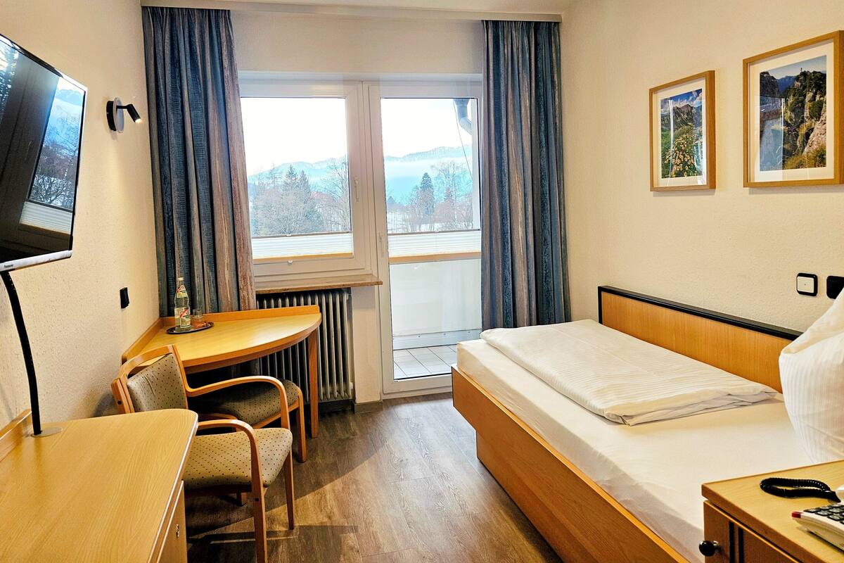 Een hotelkamer met een bed, een bureau, een stoel, een televisie, een raam met gordijnen, en twee foto's in lijsten aan de muur.