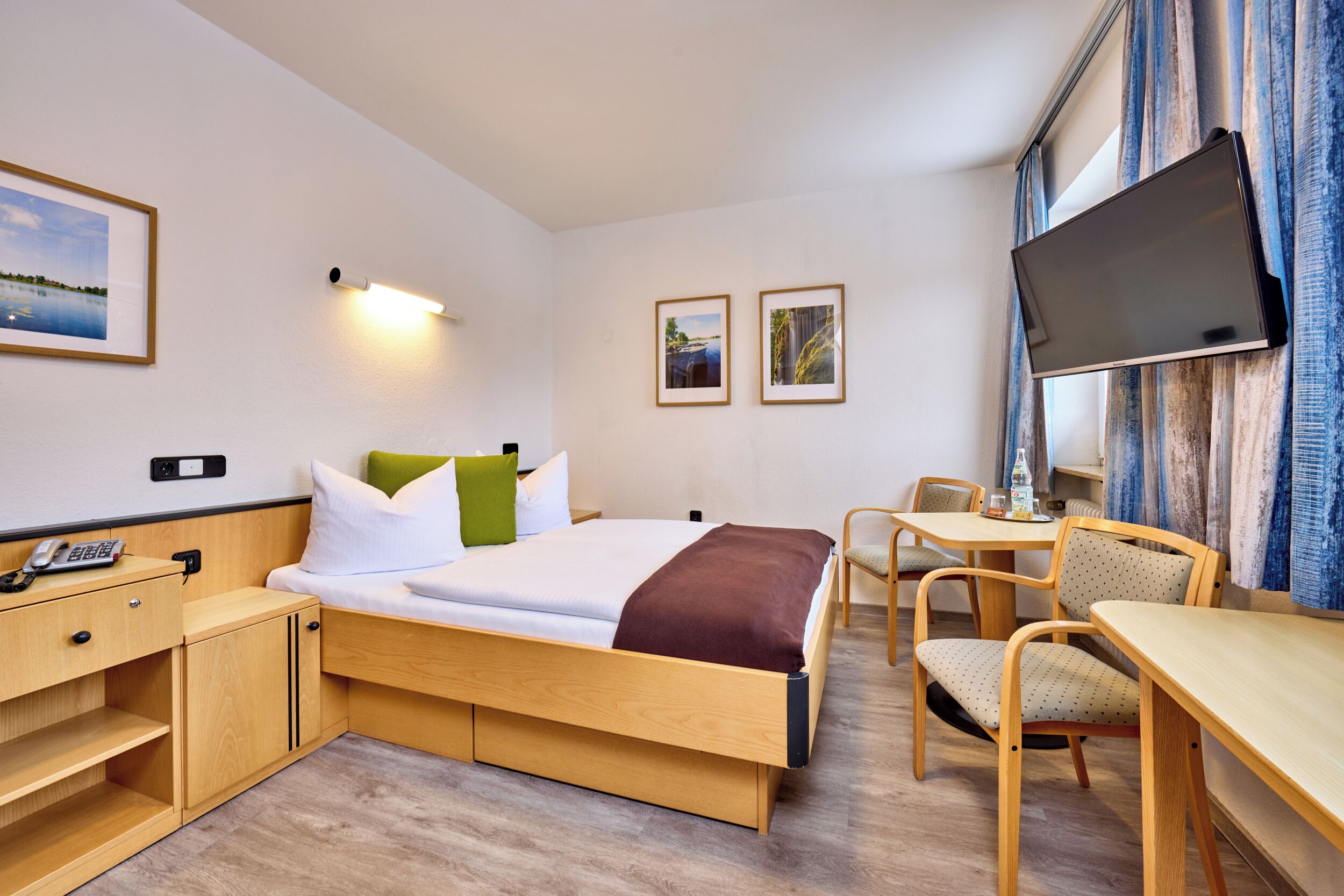 A hotel room with a bed, a desk, a chair, a TV, and artwork on the walls.