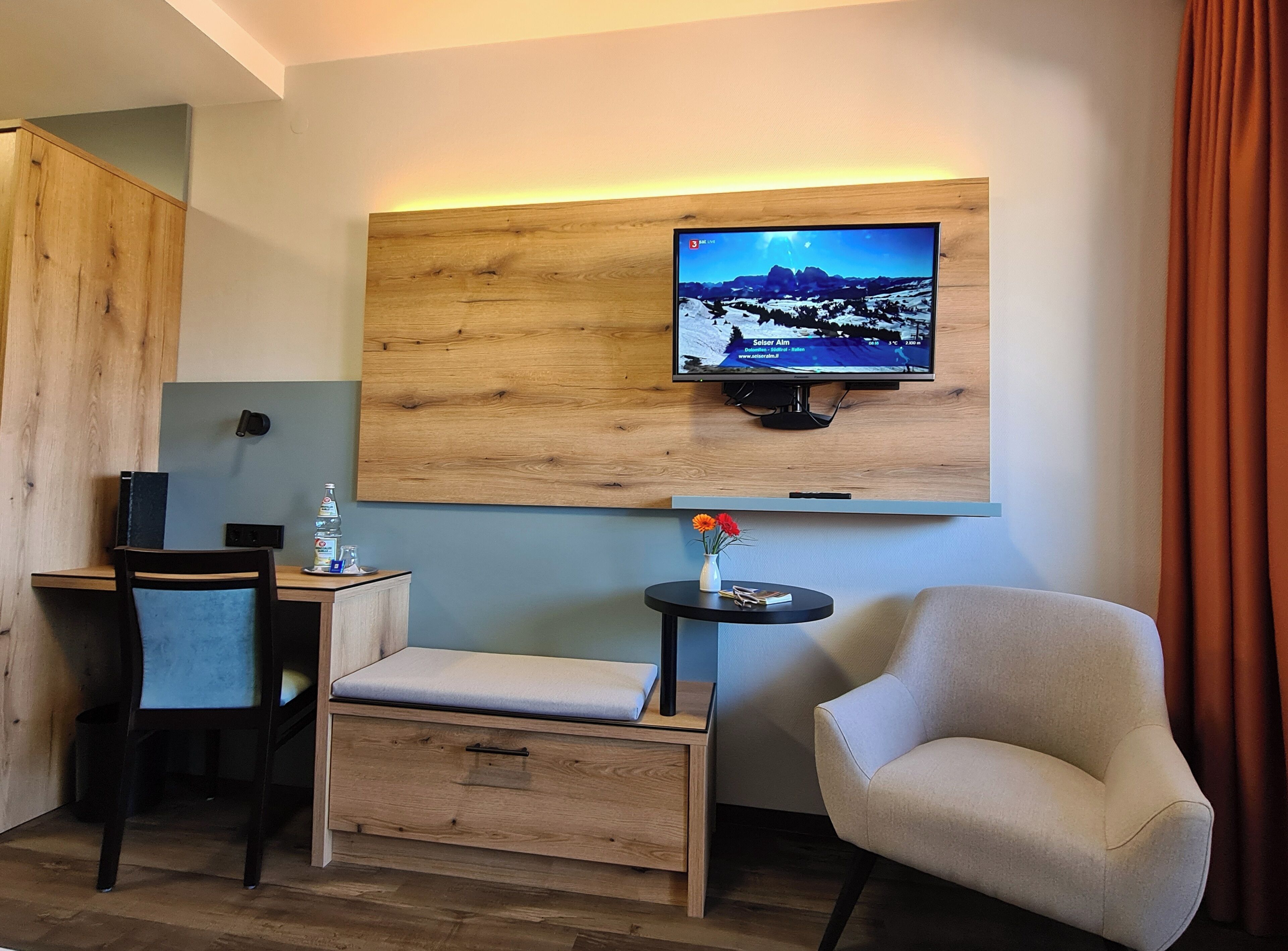 A hotel room with a wooden desk, a chair, a sofa, a television mounted on a wooden wall, and a small table with a vase of flowers.