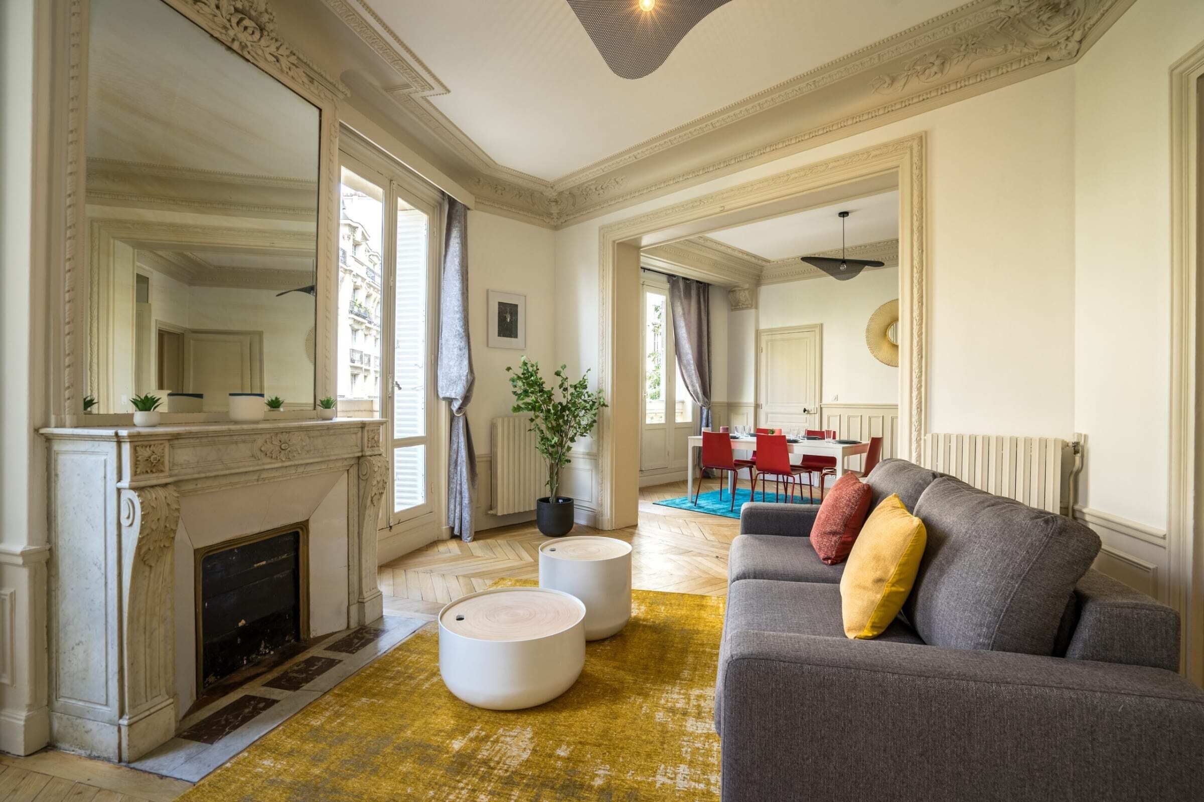 Un salon spacieux avec une cheminée, un canapé gris agrémenté de coussins, un coin repas avec des chaises rouges et un grand miroir qui reflète la pièce.