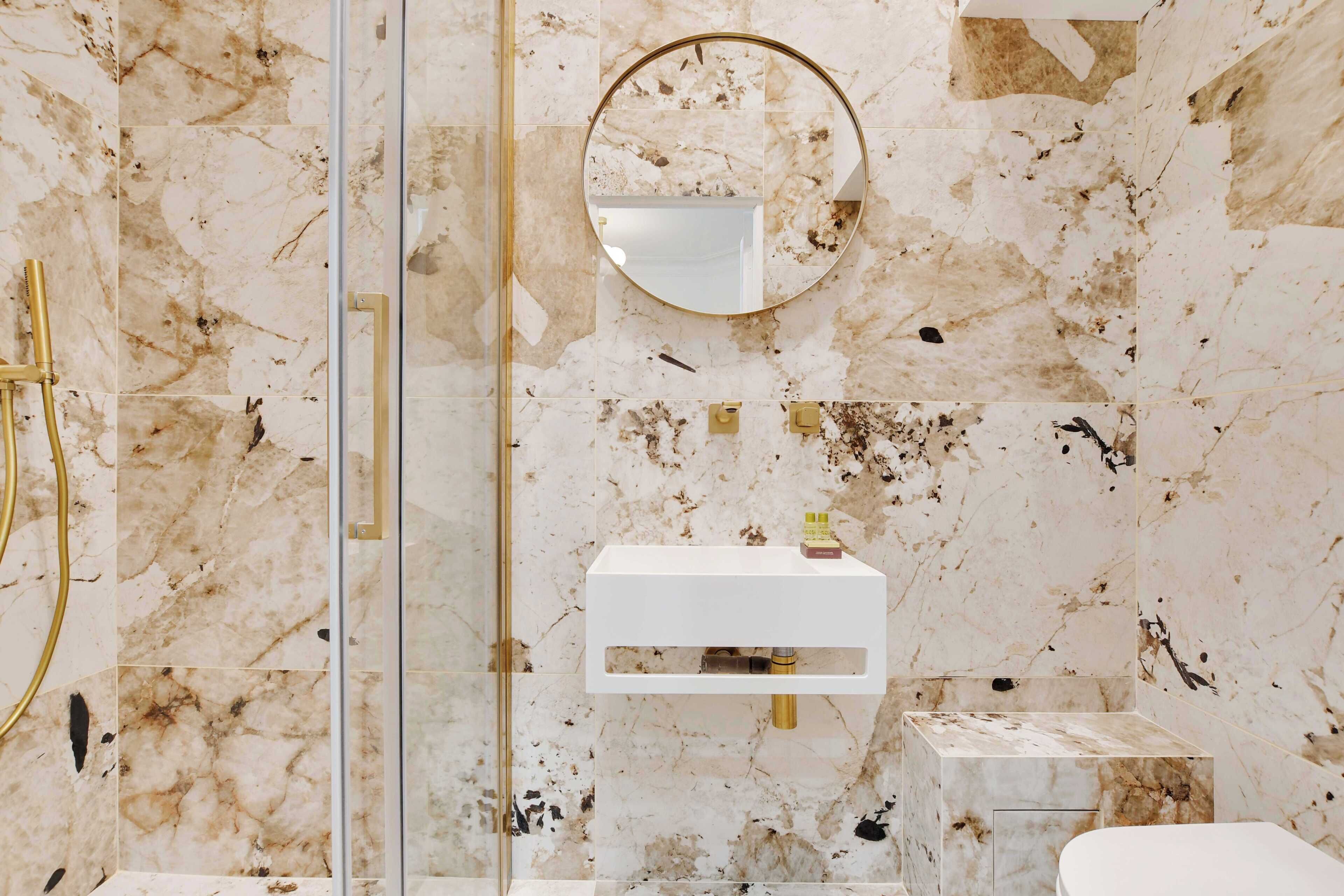 A bathroom with a marble wall, a glass shower enclosure, a white sink, and a round mirror.