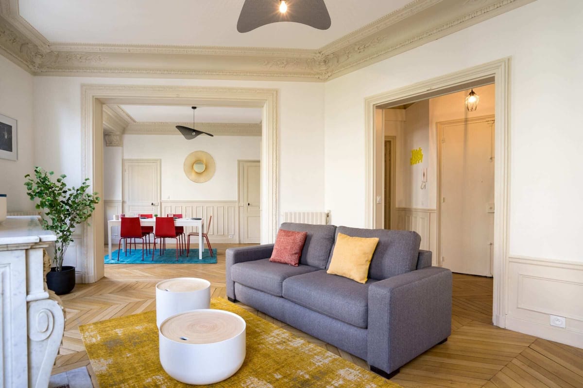 A living room with a grey sofa, a round coffee table, and a yellow rug. There is a dining area in the background with red chairs and a white table.