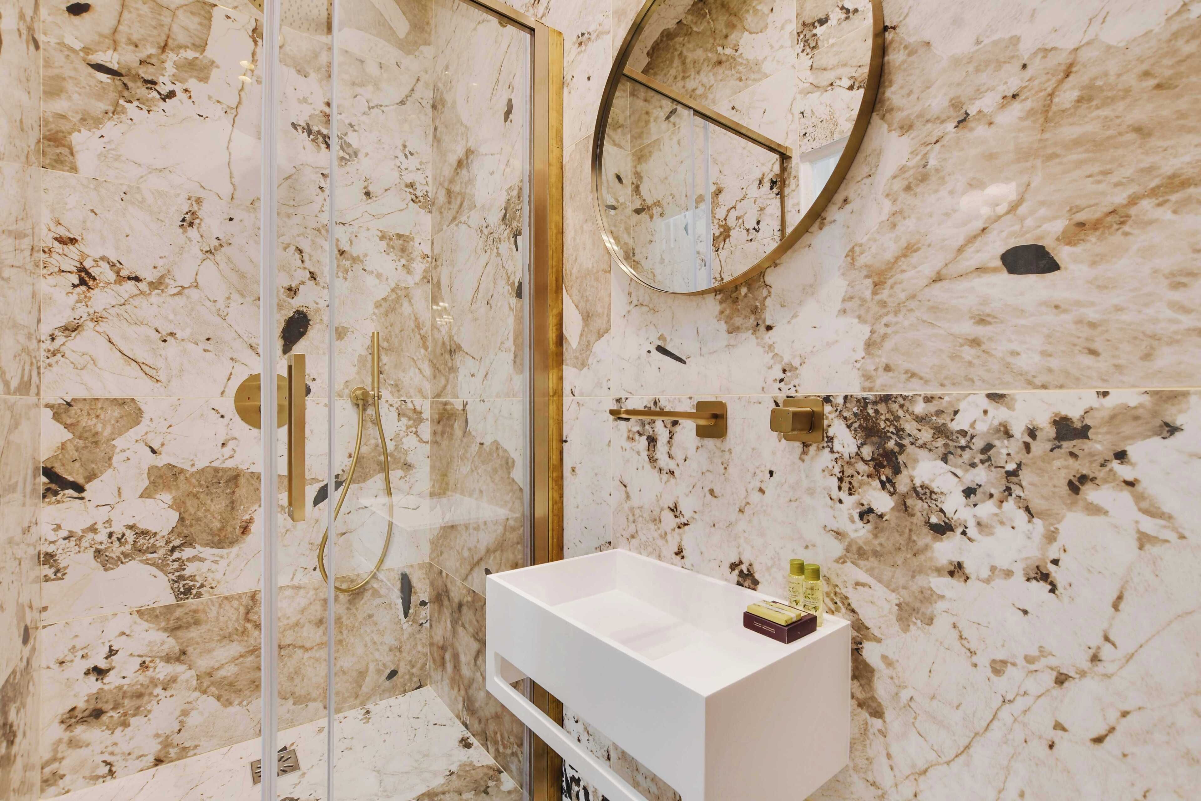 Une salle de bain avec un mur en marbre, une douche en verre, un lavabo blanc et un miroir avec un cadre doré.