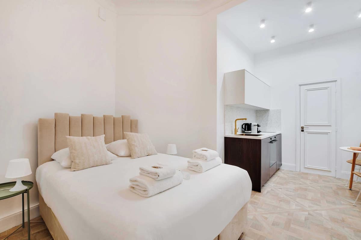 A neatly made bed with white linens and towels, a bedside table with a lamp, and a small kitchenette area with a coffee maker.