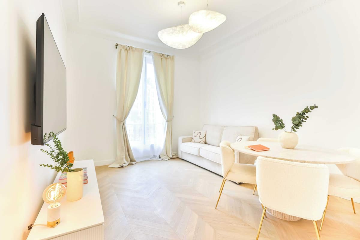 A modern living room with a sofa, dining table, and chairs, a flat-screen TV, and a large window with curtains.