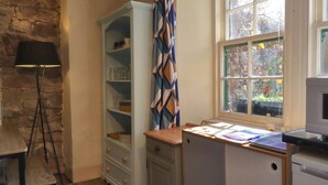 Eine Küche mit einem Holztisch, einem Schrank mit Gläsern, einem gemusterten Vorhang und einem Fenster mit Blick auf Grünflächen.