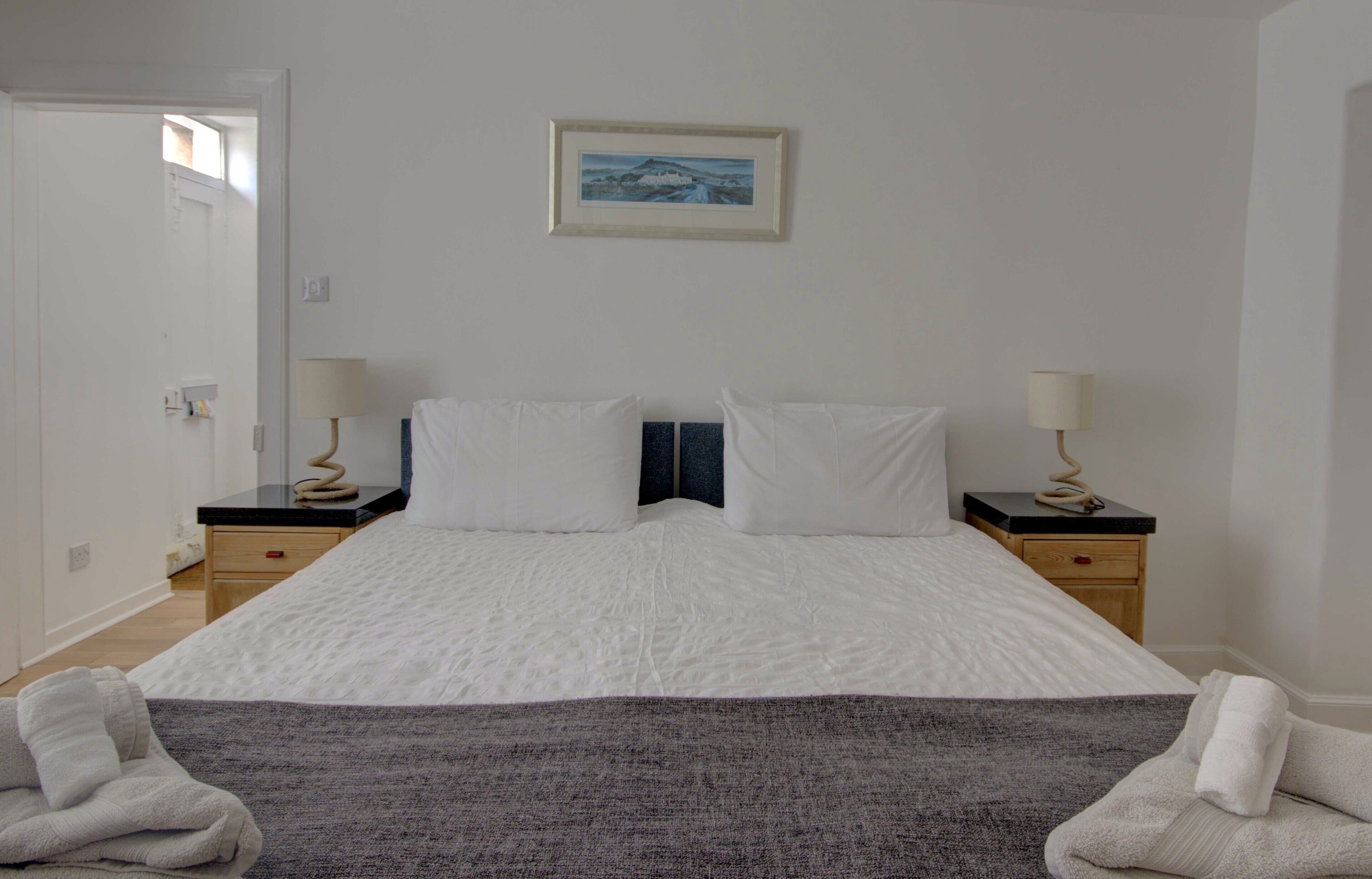 Un letto matrimoniale rifatto con lenzuola bianche, due comodini con lampade, un quadro incorniciato appeso al muro e una coperta grigia sul letto.