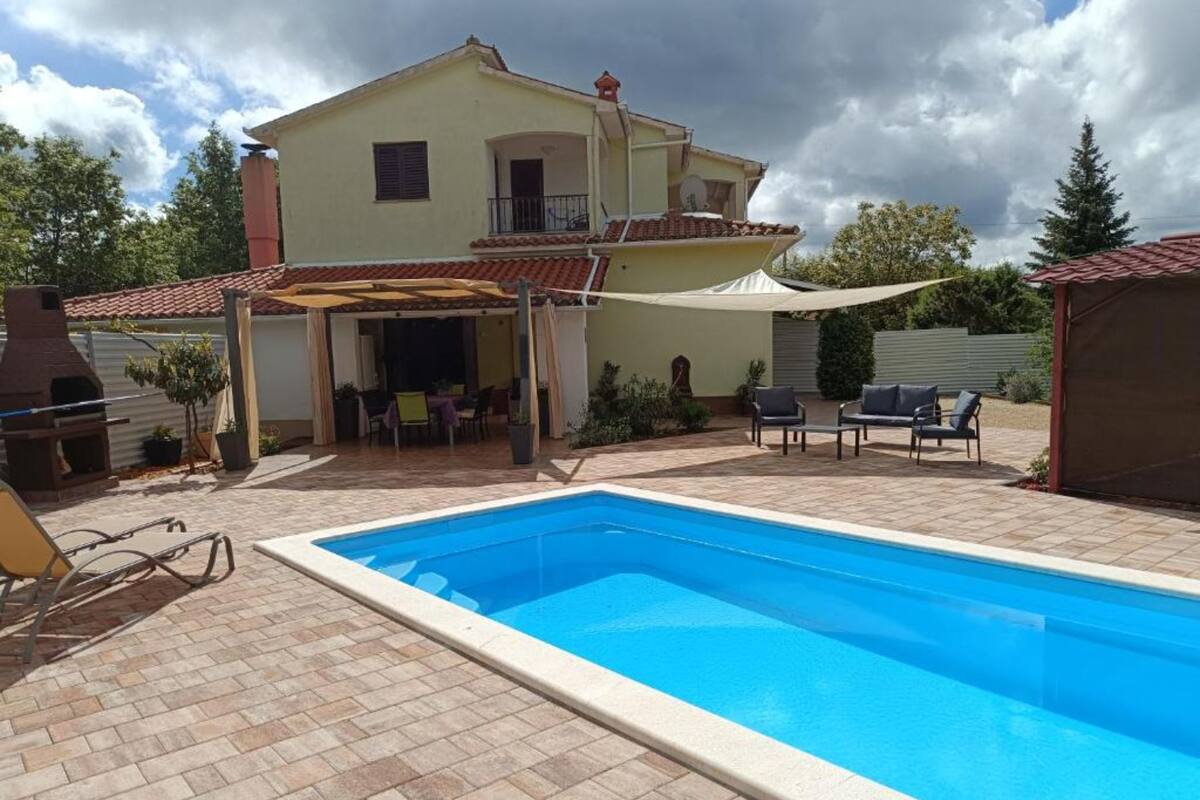 A swimming pool with a tiled deck, a covered patio area, and a house with a red-tiled roof.