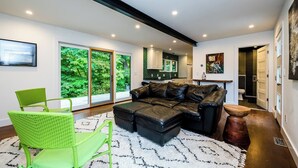 A modern living room with a black leather sectional sofa, a green wicker chair, and a white patterned rug. - Unique container home retreat near Lake Michigan. Enjoy a peaceful, private setting with a spacious deck, firepit, and ravine views. Close to beaches, wineries, and downtown St. Joseph—perfect for a relaxing Southwest Michigan getaway. (Benton Harbor)
