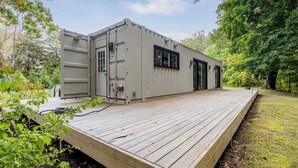 A compact, grey, container-style building with a wooden deck, large windows, and a door. - Unique container home retreat near Lake Michigan. Enjoy a peaceful, private setting with a spacious deck, firepit, and ravine views. Close to beaches, wineries, and downtown St. Joseph—perfect for a relaxing Southwest Michigan getaway. (Benton Harbor)
