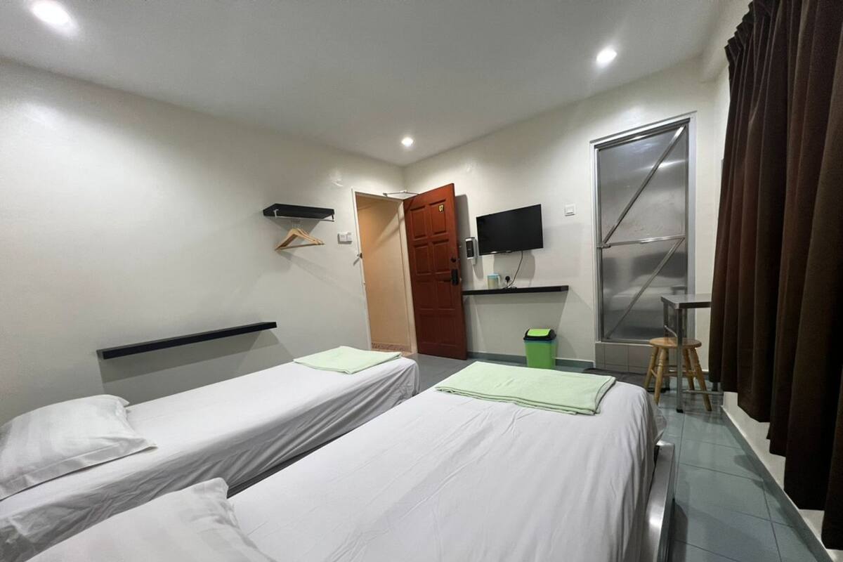 Habitación de hotel con dos camas individuales, televisión, una mesita, una silla, ventana con cortinas y una puerta.