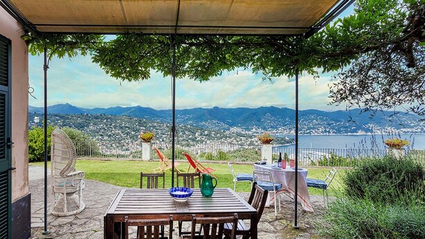 A patio with a table and chairs, a view of a city and mountains, and a body of water in the background.