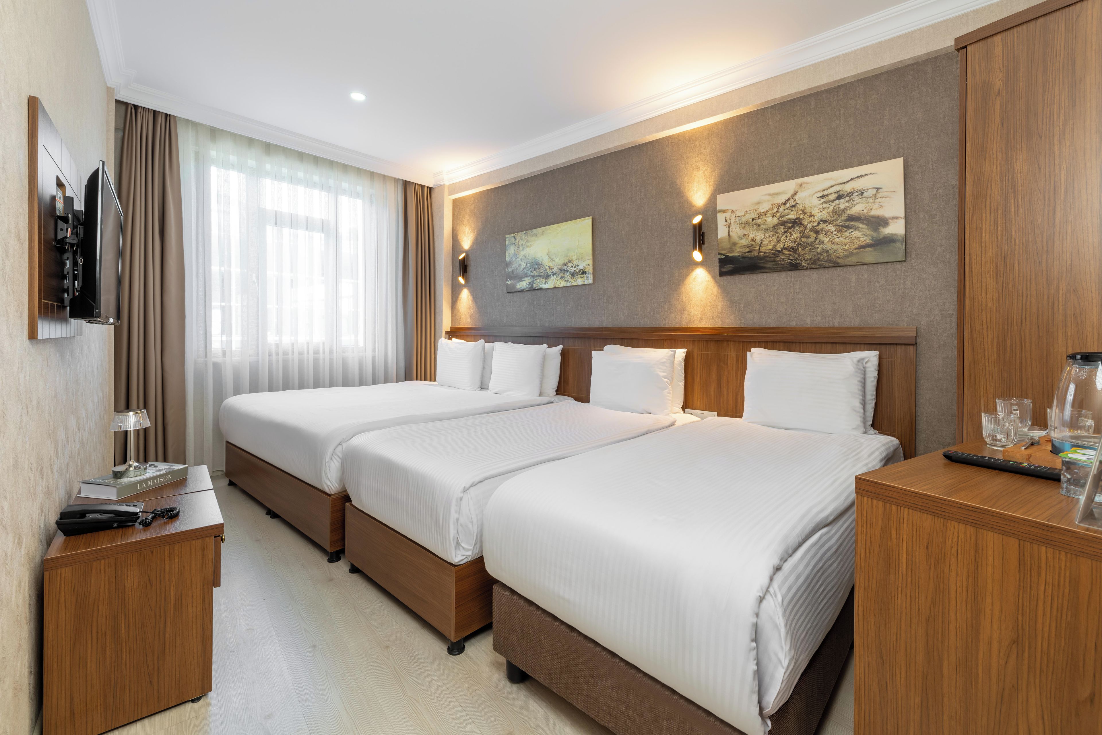 A hotel room with two beds, a flat-screen TV mounted on the wall, a bedside table with a lamp, a remote control, and a glass of water.