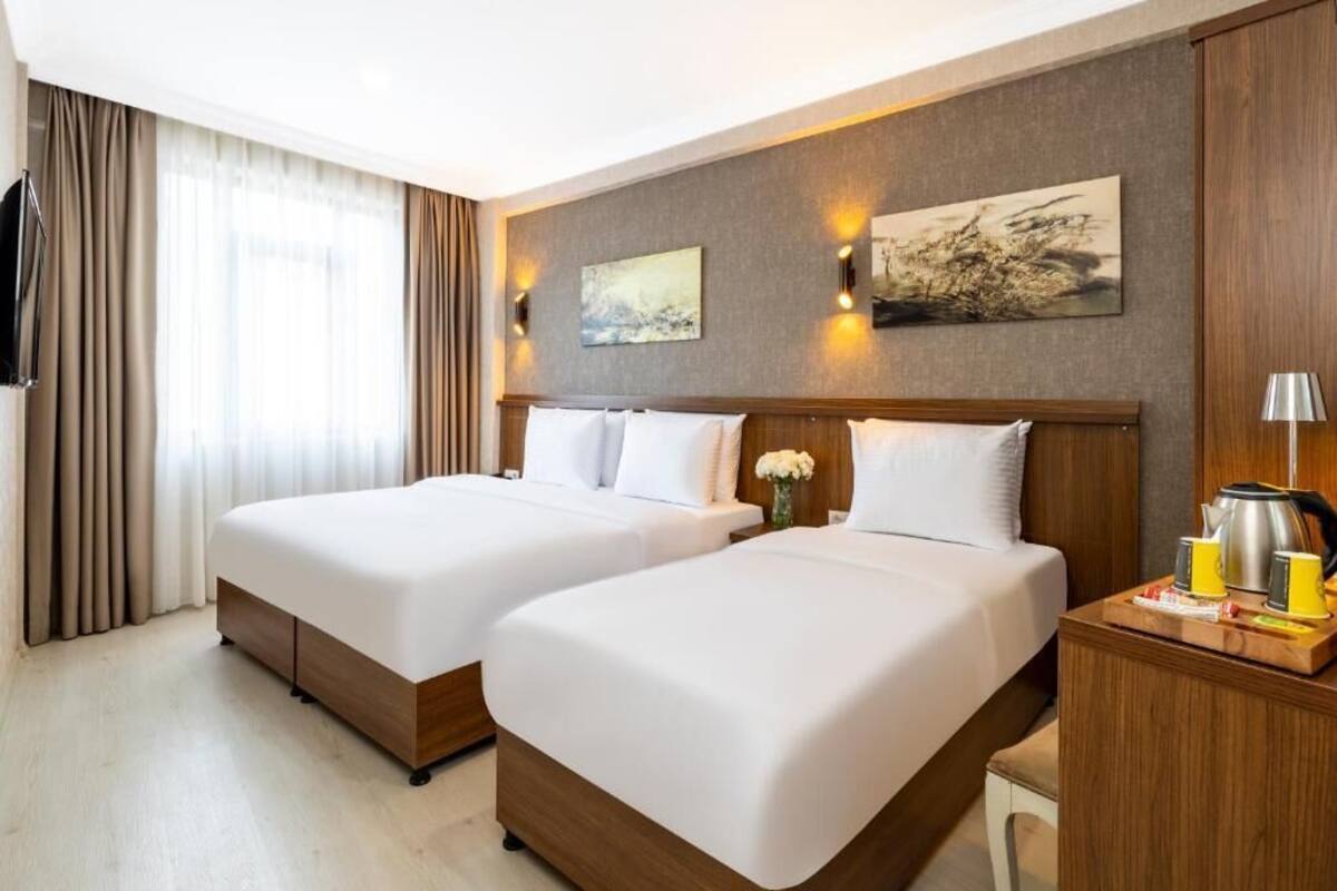 A hotel room with two beds, a wooden headboard, a flat-screen TV, a painting, and a bedside table with a kettle and cups.