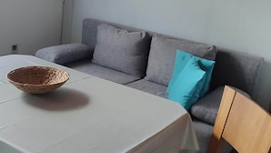 Une pièce avec un canapé gris, une chaise en bois, une table recouverte d’une nappe blanche et un tableau accroché au mur.