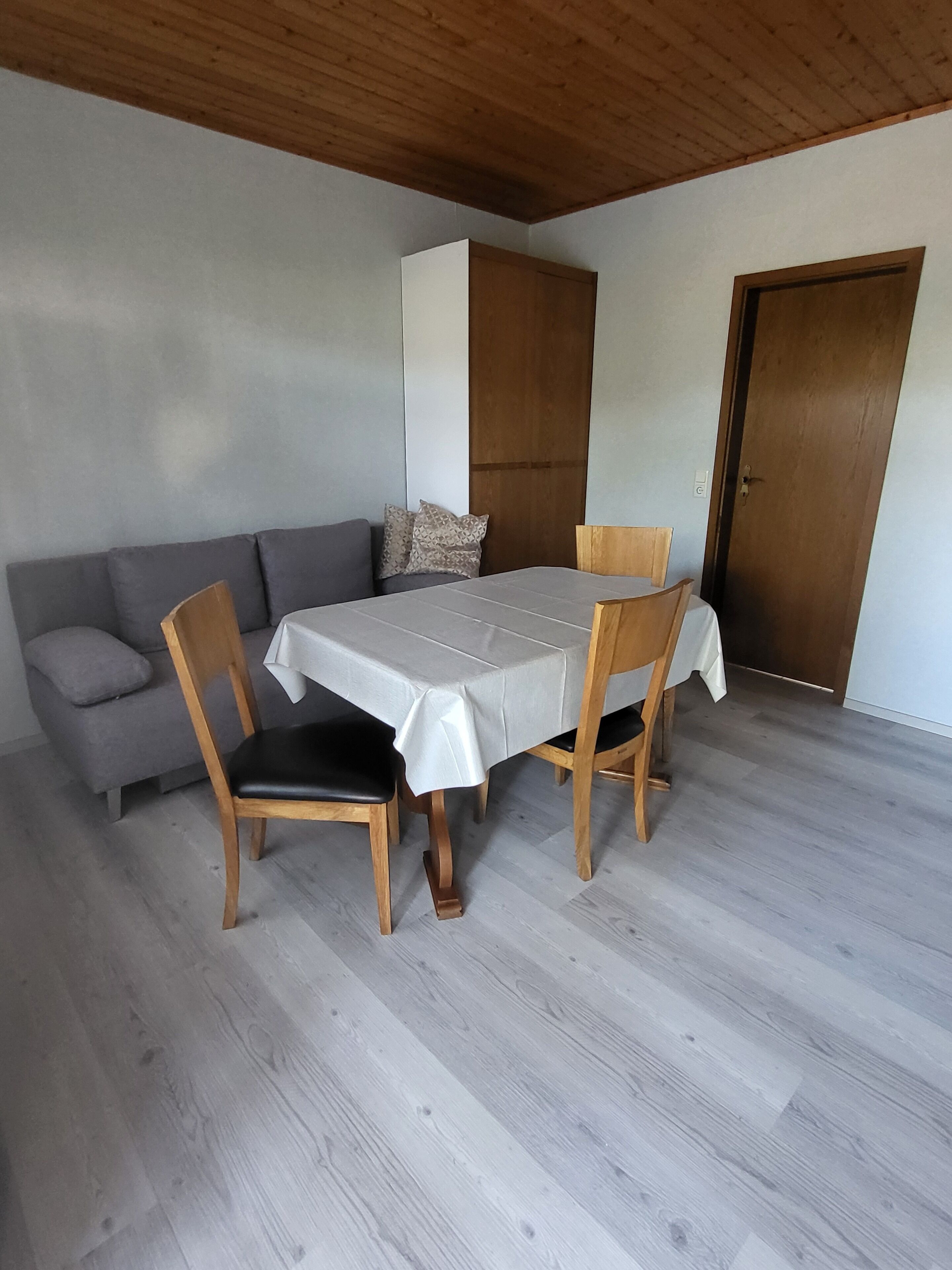 Zona pranzo con tavolo e quattro sedie, un divano grigio con cuscini e una porta in legno.