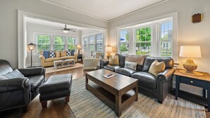 A spacious living room with a leather sofa, a coffee table, and a side table with a lamp. There are multiple windows providing natural light and a view of the outdoors. - Cozy Corner Cottage| Beach Access & Historic Charm (Sheboygan)
