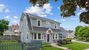 A two-story house with a front porch, a picket fence, and a well-maintained lawn. - Cozy Corner Cottage| Beach Access & Historic Charm (Sheboygan)