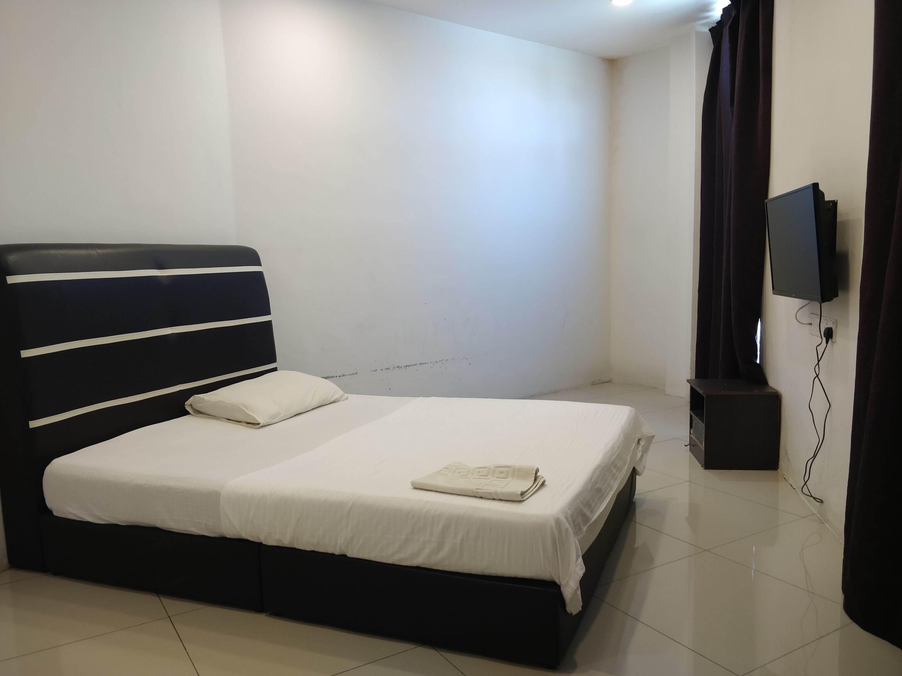 A neatly made bed with white linens and a folded towel, a black headboard with white stripes, a flat-screen TV mounted on the wall, and a dark-colored speaker.