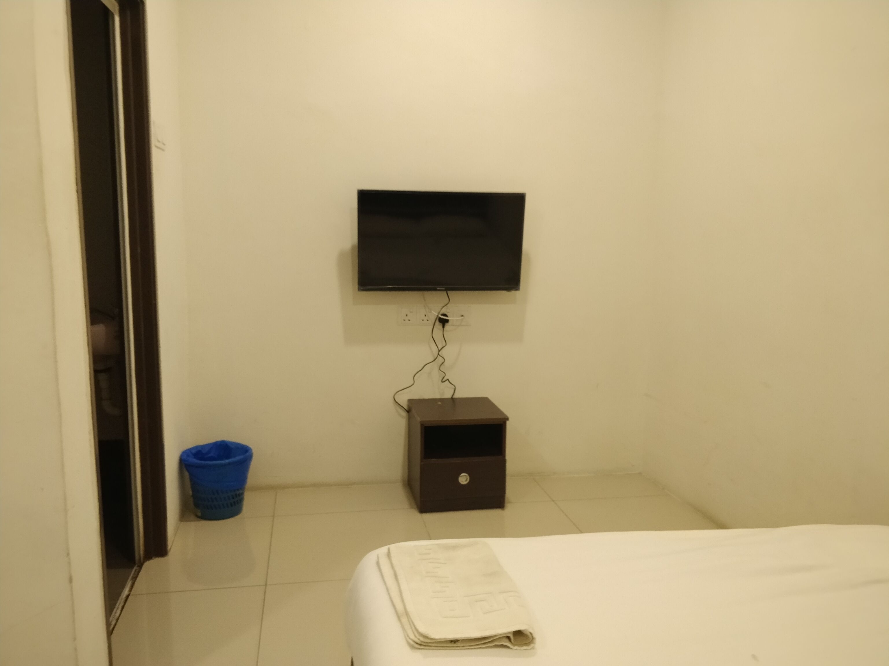 A hotel room with a bed, a television mounted on the wall, a small drawer unit, and a blue laundry basket.