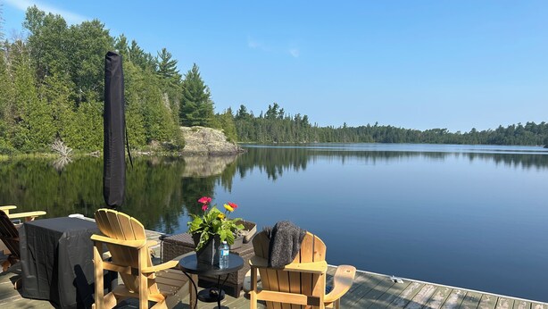 A wooden deck with outdoor seating and a table, overlooking a calm lake with trees and a distant island. - Granite Lake Chalet - waterfront Temagami/Hot Tub (Temagami)