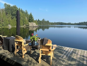 A wooden deck with outdoor seating and a table, overlooking a calm lake with trees and a distant island. - Granite Lake Chalet - waterfront Temagami/Hot Tub (Temagami)