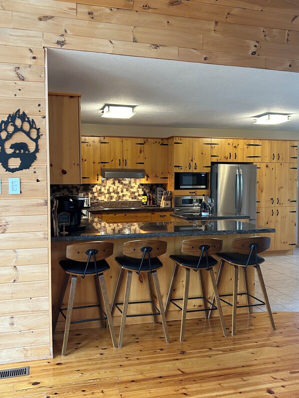 A modern kitchen with wooden cabinets, a stainless steel refrigerator, and a bar area with stools. - Granite Lake Chalet - waterfront Temagami/Hot Tub (Temagami)