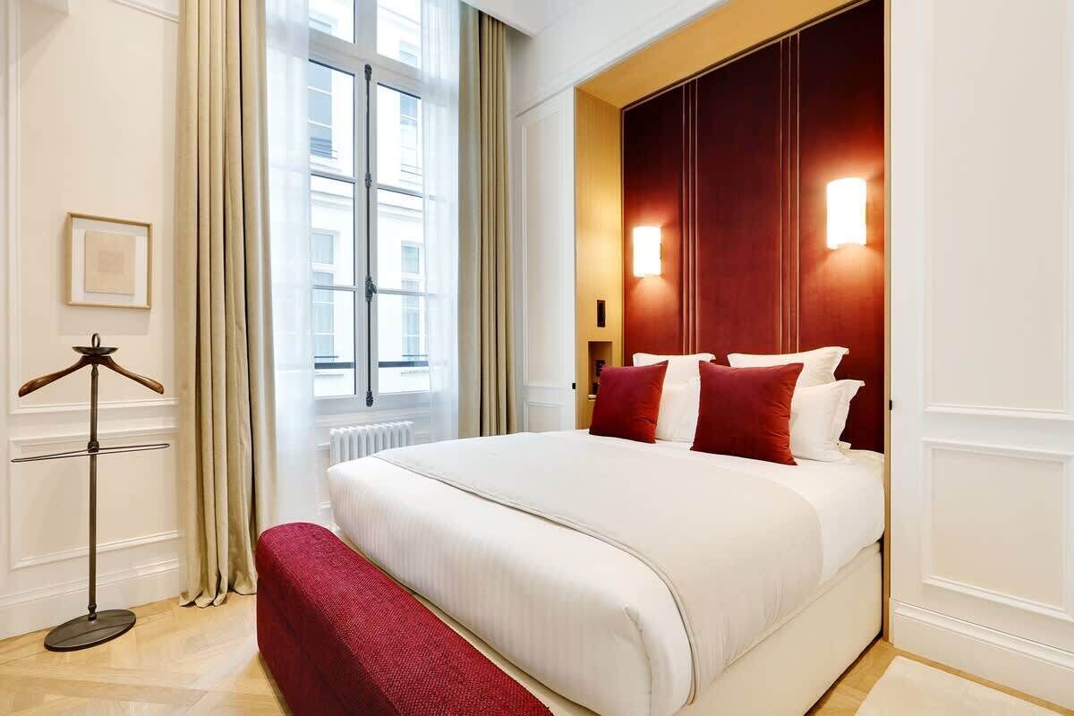 Una habitación de hotel con una cama grande, un banco rojo, una lámpara de pie y una ventana con cortinas.