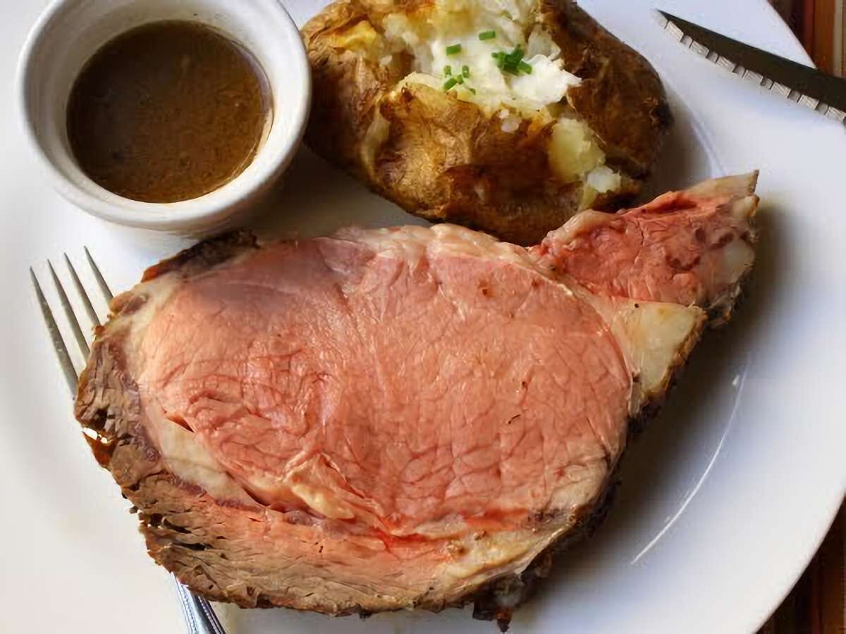 A plate with a slice of roast beef, a baked potato, and a small bowl of gravy.