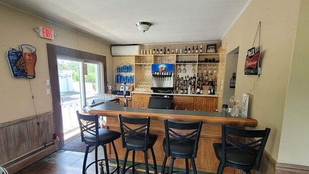 A bar area with a wooden counter, black bar stools, and a variety of bottles and glasses on shelves. - DuBois' Inn (Madawaska)