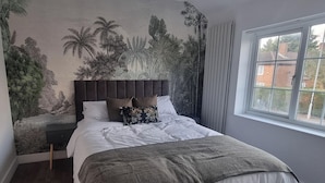 Ein Schlafzimmer mit einem großen Bett, einem Kopfteil, einem Nachttisch, einem Fenster mit Vorhängen und einem Wandgemälde mit tropischen Pflanzen.