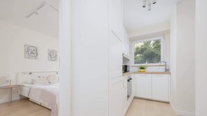 A modern, minimalist bedroom and kitchen with white cabinetry, a wooden floor, and a large window providing natural light. - Bright Apartment with Balcony | Workplace, Close to Metro (Warszawa)