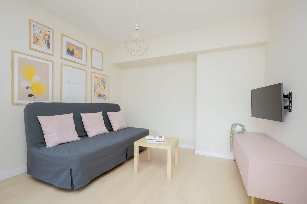 Una sala de estar moderna con un sofá gris, una mesita de madera, un banco rosa y un televisor montado en la pared.