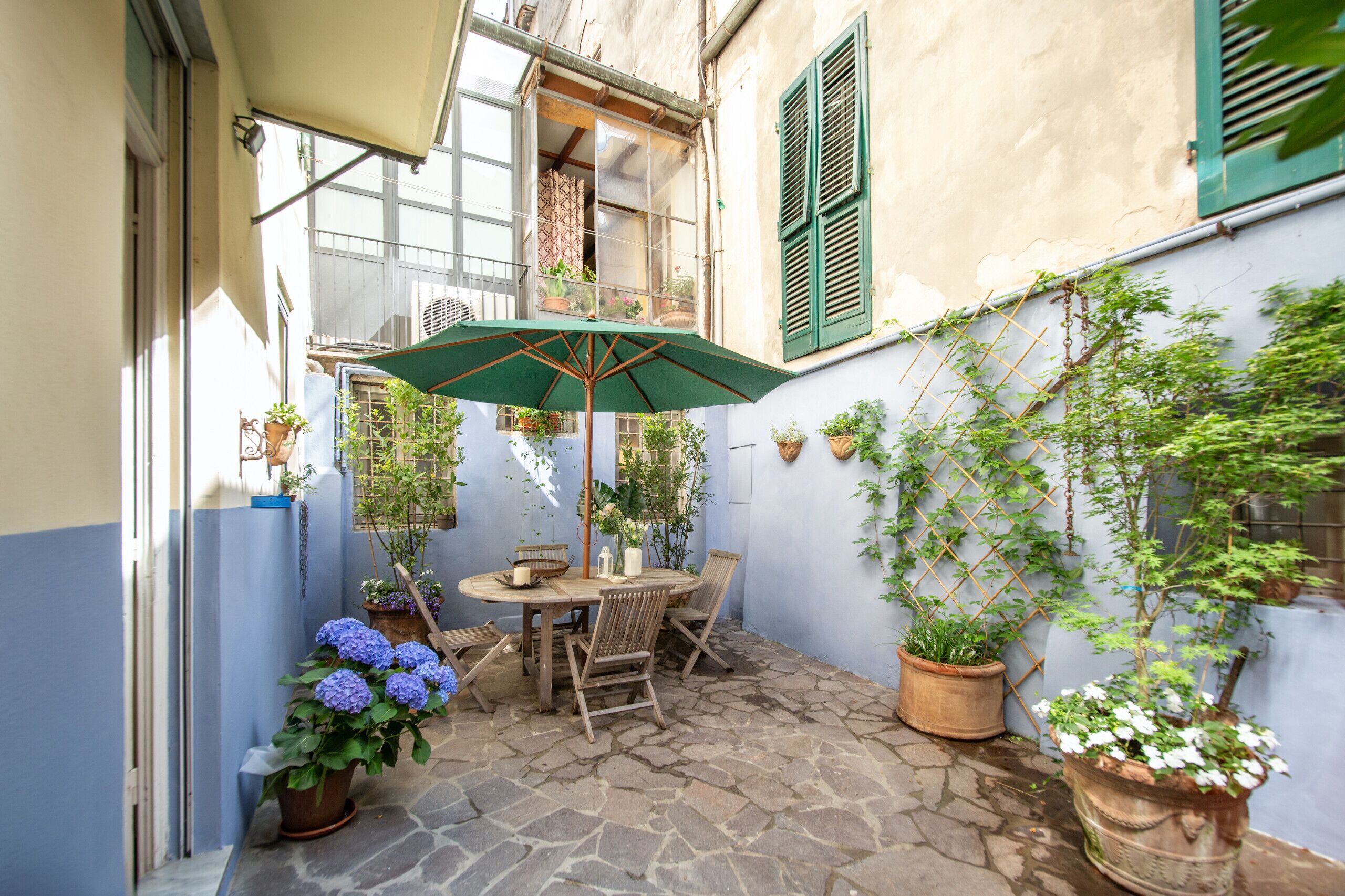 Eine Terrasse mit Steinboden, einem Holztisch und -stühlen, umgeben von Topfpflanzen und einem grünen Sonnenschirm.