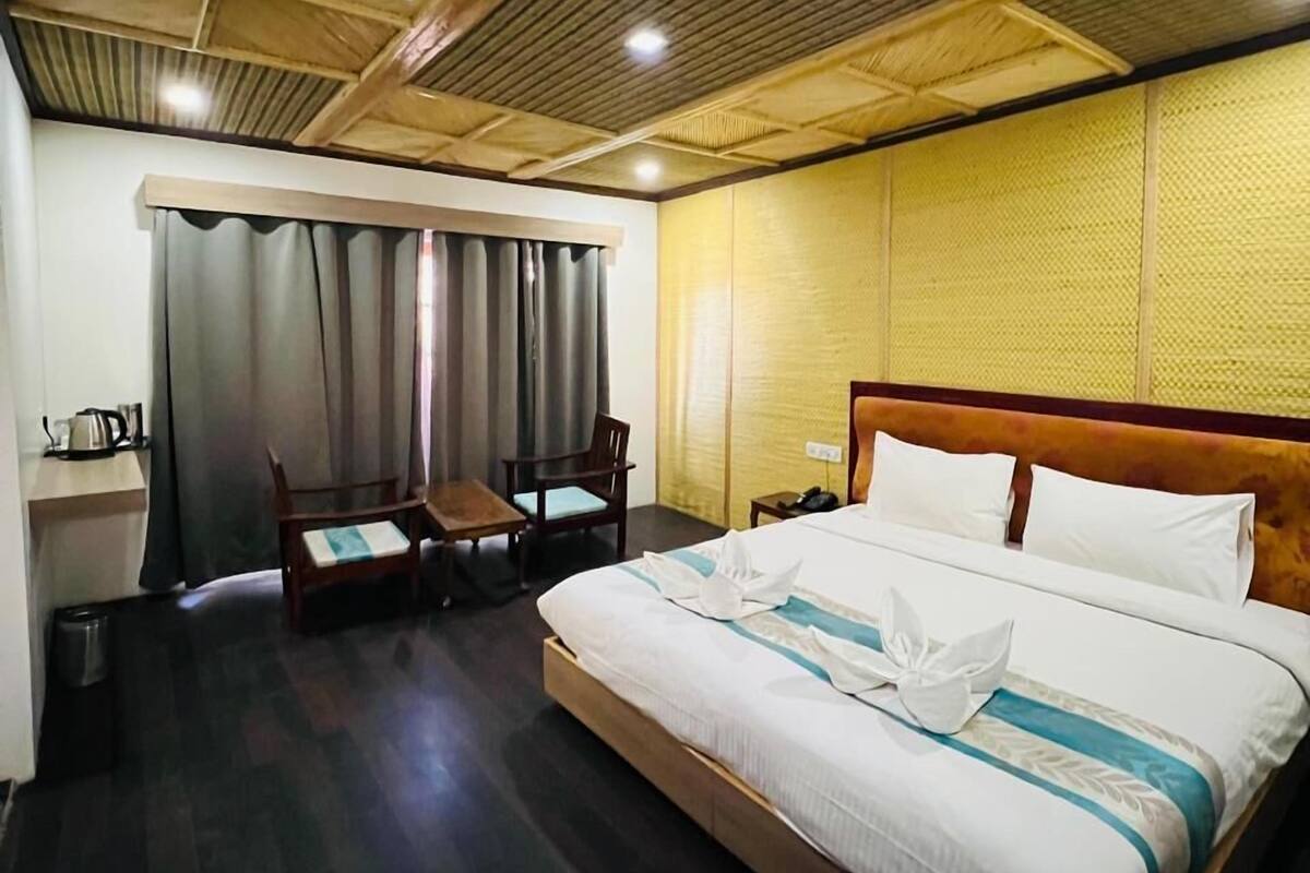 A hotel room with a wooden ceiling, a bed with white bedding, a wooden nightstand, a chair, a desk with a kettle, and a trash bin.