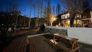 An outdoor patio area with wooden benches and a stone wall, surrounded by trees and a building with illuminated windows. - Thongsal (Leh)