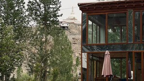 A modern building with large windows and a patio area with white chairs and a pink umbrella. - Thongsal (Leh)