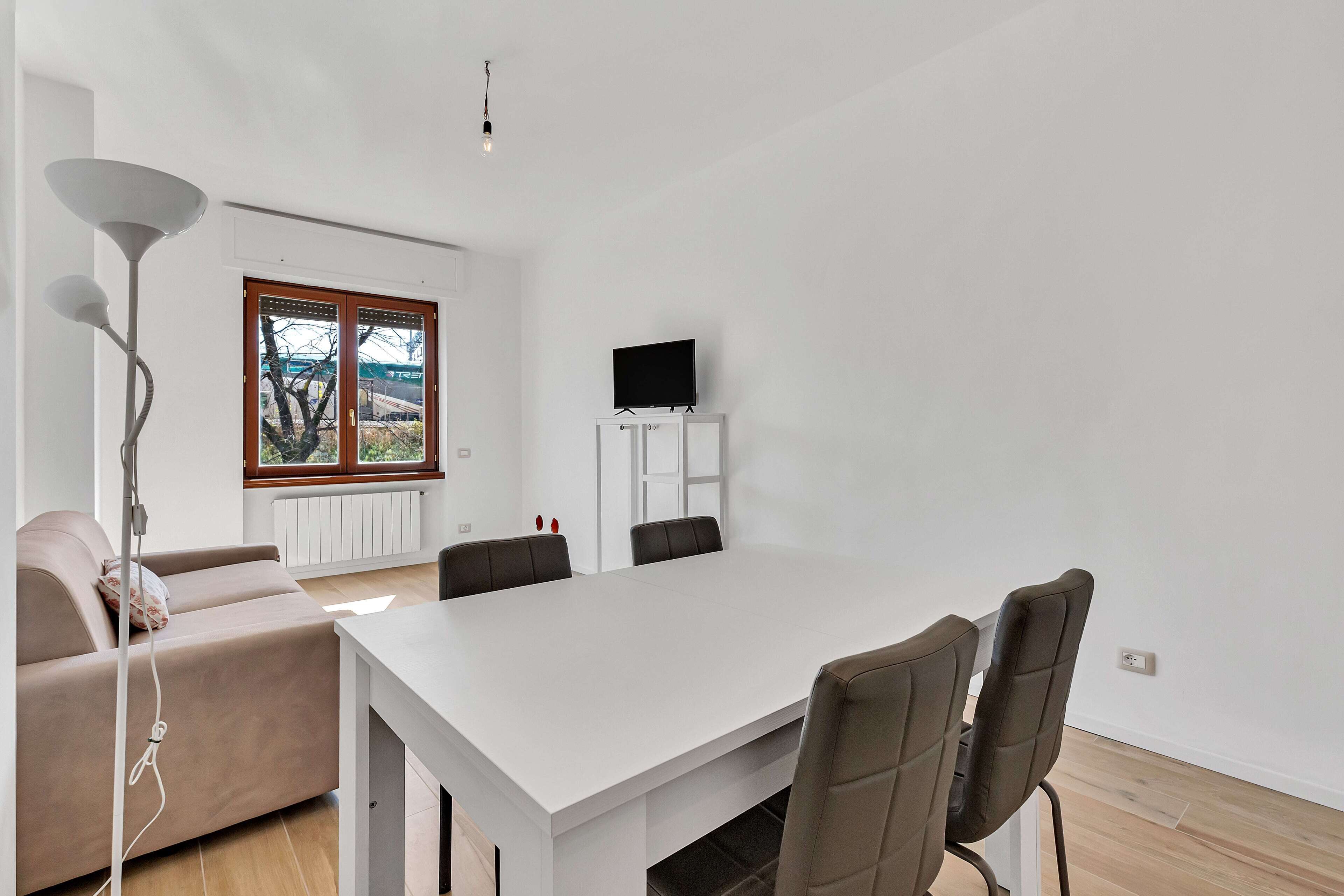 Una sala moderna con un sofá blanco, una mesa de comedor con sillas, un televisor sobre un mueble bajo y una lámpara de pie.