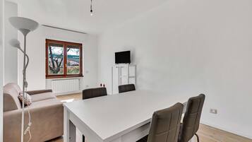 Una sala moderna con un sofá blanco, una mesa de comedor con sillas, un televisor sobre un mueble bajo y una lámpara de pie.