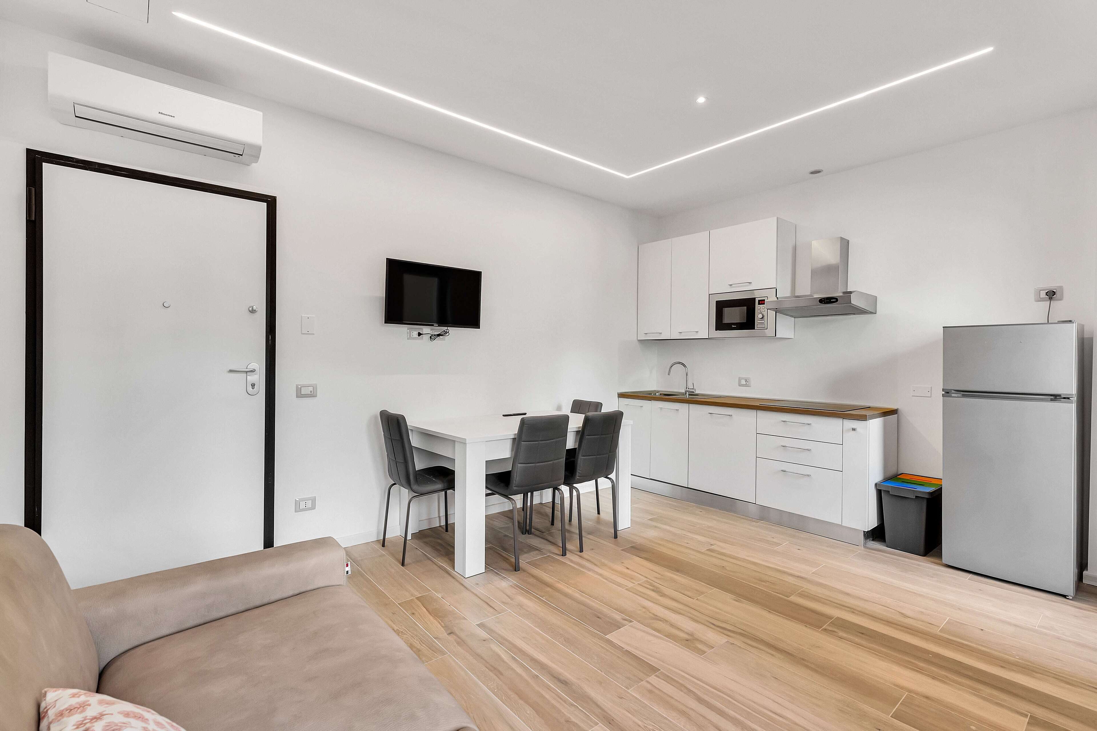 A modern kitchen with white cabinets, a microwave, and a refrigerator. There is a dining table with chairs and a TV mounted on the wall.