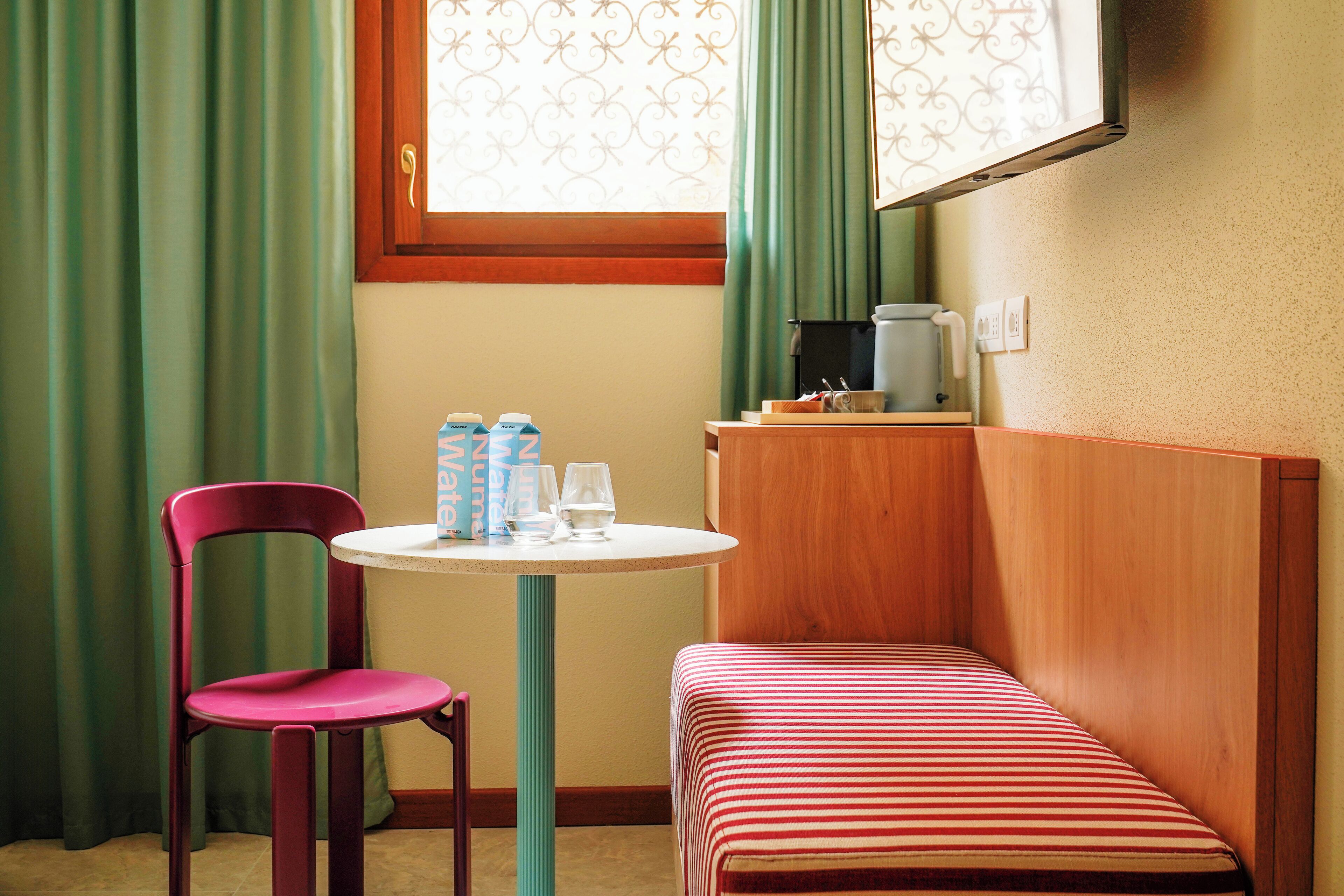 Une petite pièce avec un lit simple, une table et une chaise, une fenêtre avec des rideaux et une machine à café posée sur une étagère.