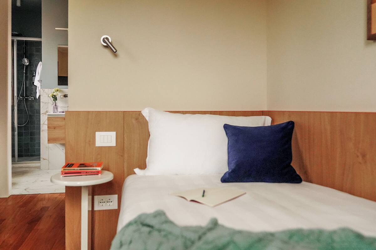 A hotel room with a wooden headboard, a bedside table with a book and a pen, a green blanket, and a shower visible in the background.