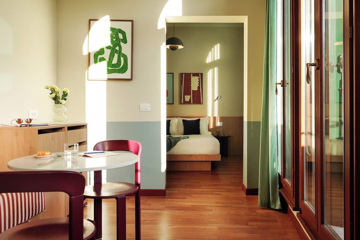 A hotel room with a bed, a desk with a chair, a small table with a chair, and a window with curtains.