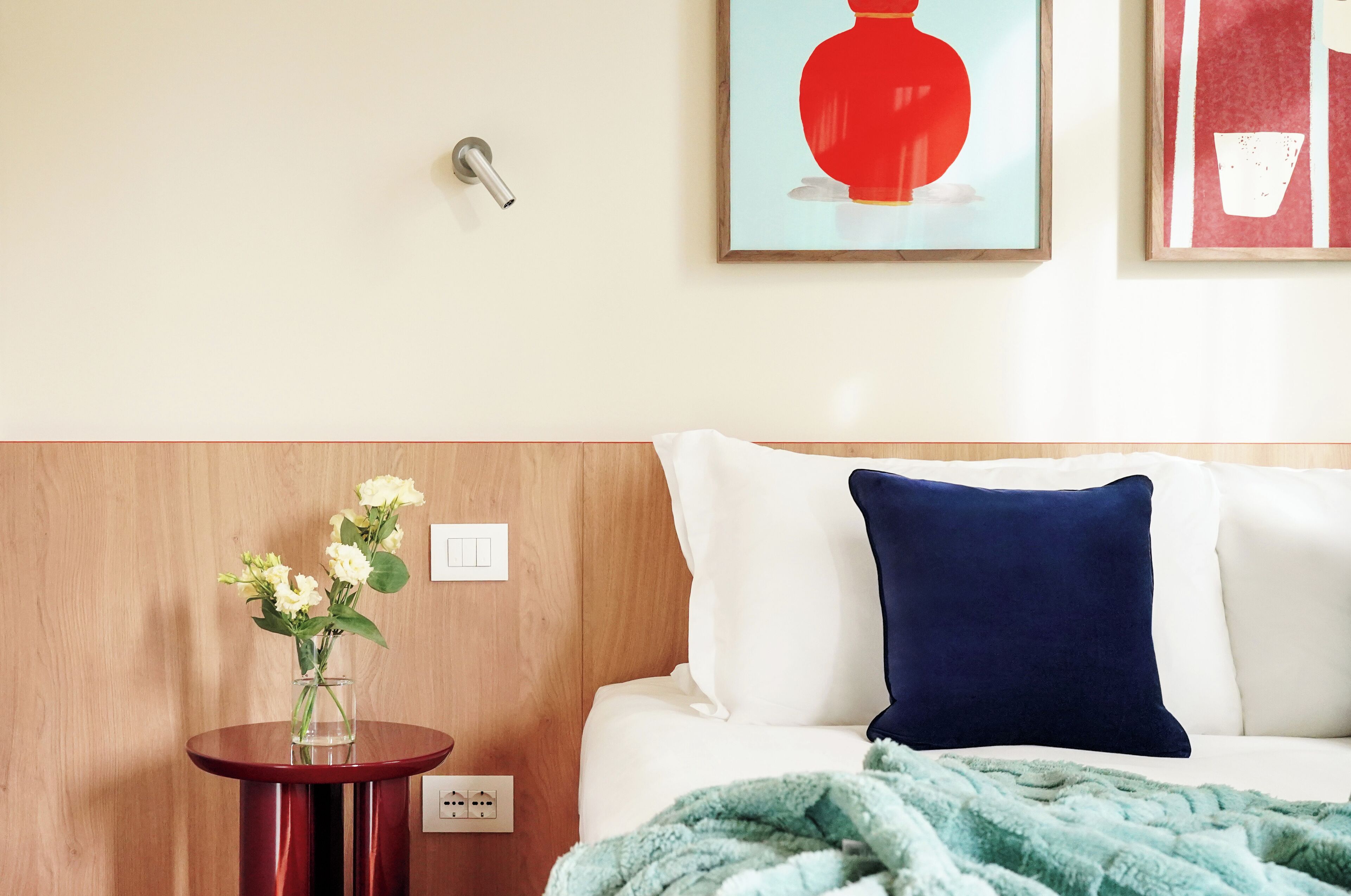 A hotel room with a wooden headboard, a white bed with a navy blue pillow, a round wooden side table with a vase of flowers, and two framed artworks on the wall.