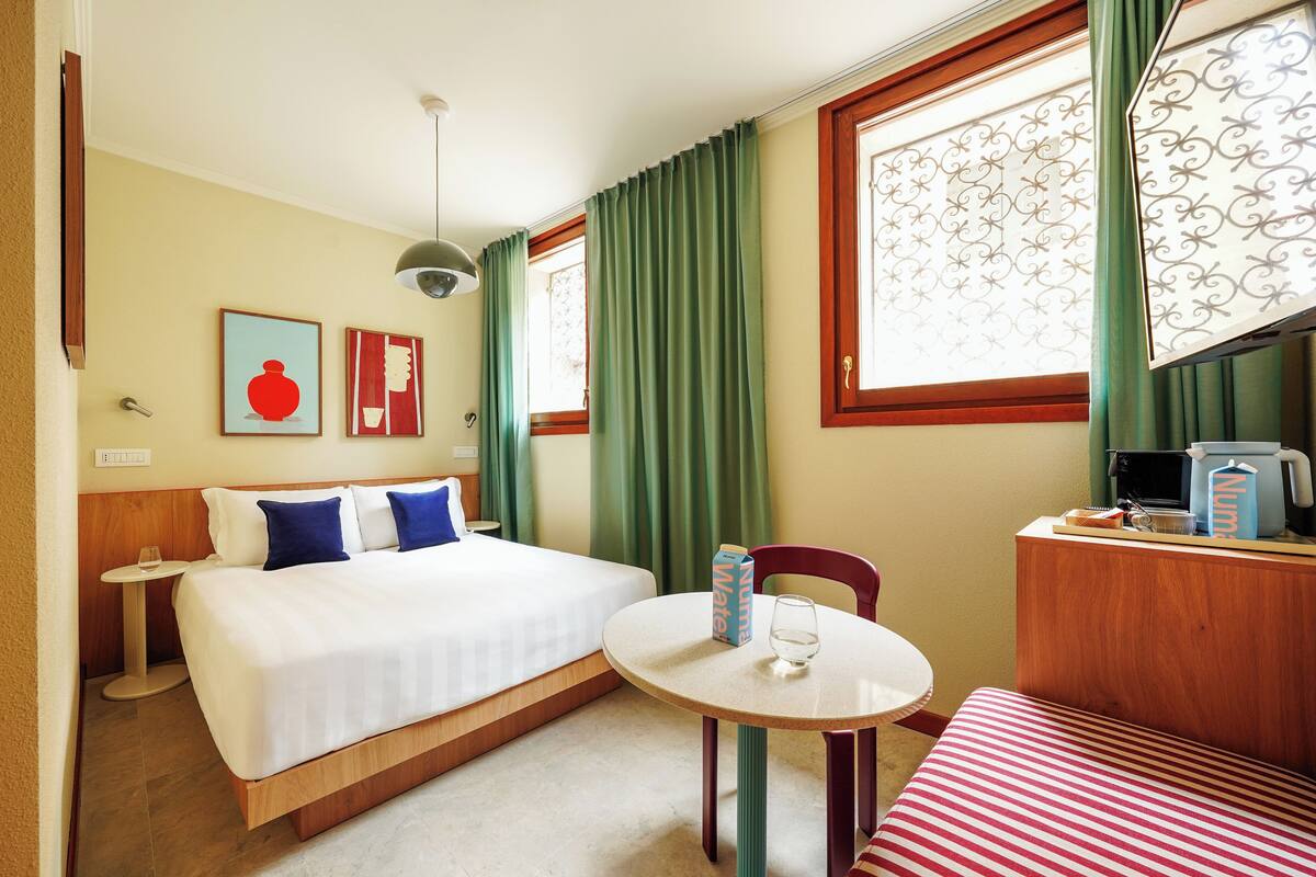 A hotel room with a bed, a round table, a chair, a small table with a glass, and a coffee machine.