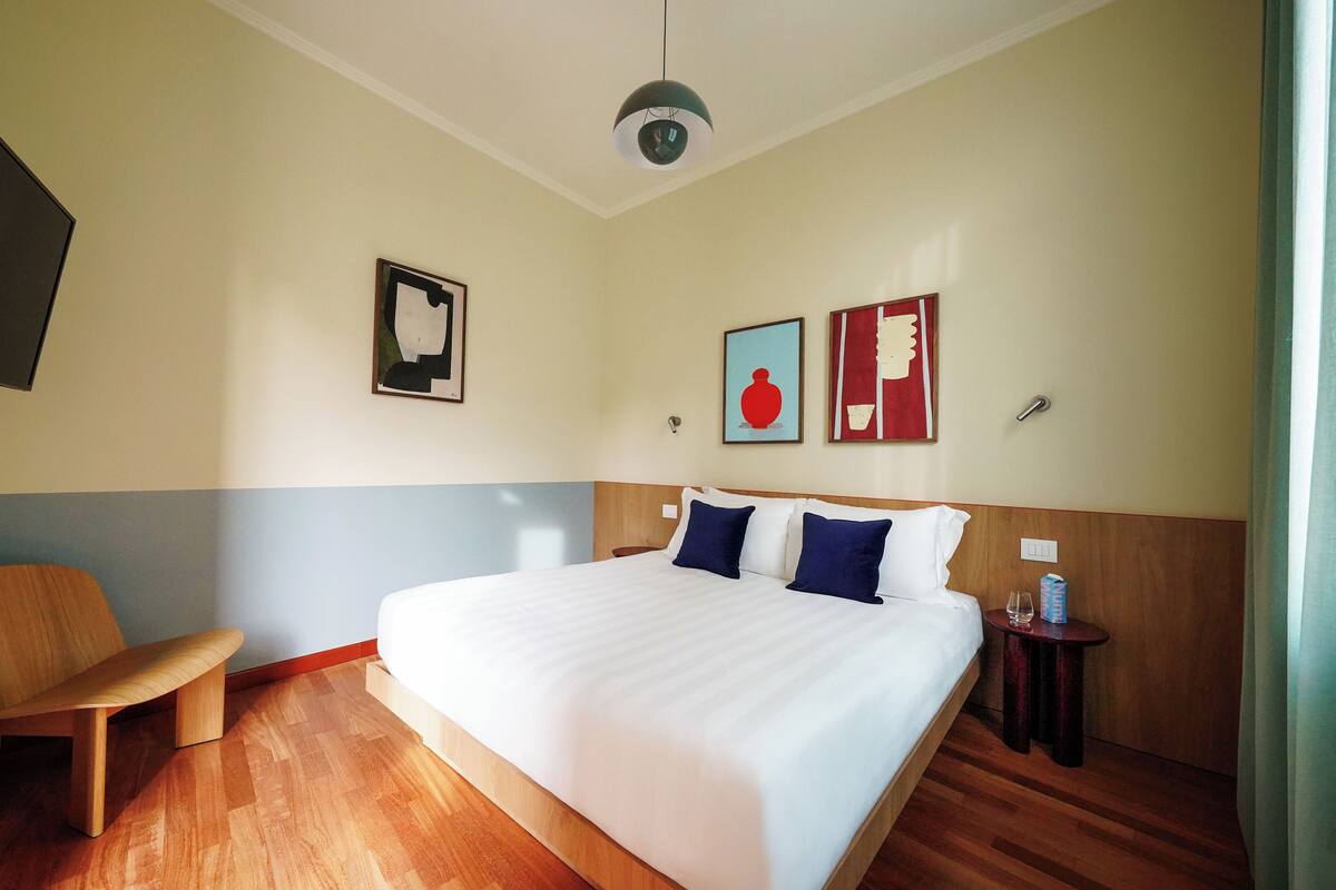 A hotel room with a large bed, a wooden bench, a small table with a glass, and artwork on the walls.