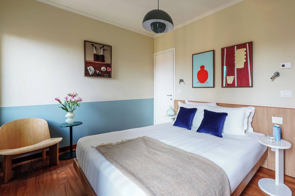 A hotel room with a large bed, a wooden chair, a small table with a vase of flowers, and two wall art pieces.