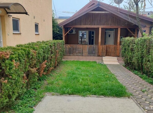 A small wooden house with a porch, a grassy yard, and a paved pathway.