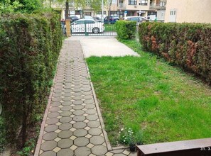 A paved pathway leading to a gated entrance, flanked by hedges and a grassy area.